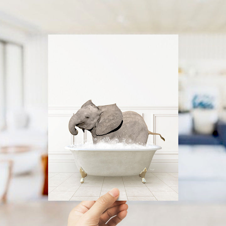 A hand holds up a framed image of a baby elephant sitting in a bathtub filled with water.