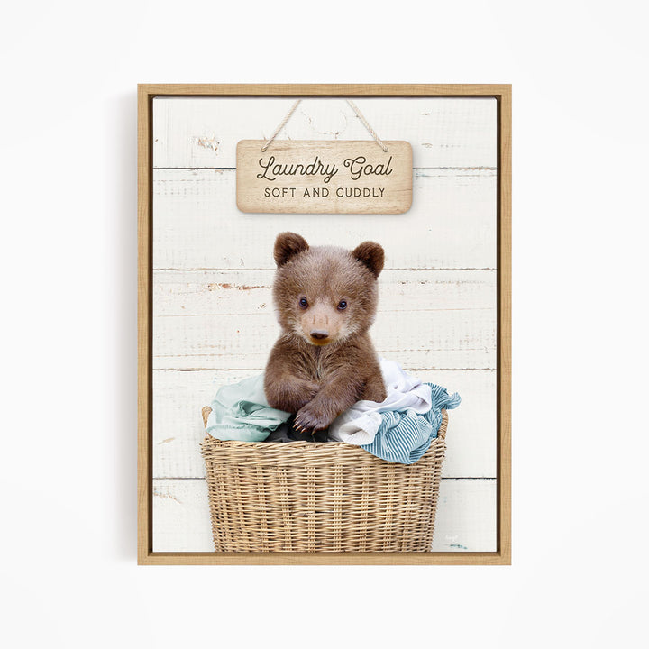 A cute brown bear sitting in a wicker laundry basket, with a wooden sign above that says "Laundry Goal Soft and Cuddly".
