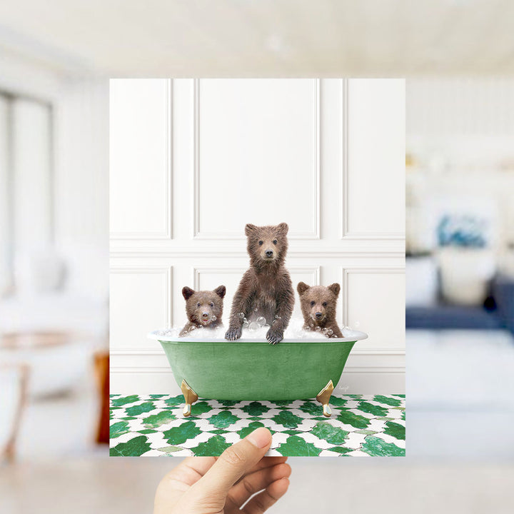 A hand holds up a framed photograph of three adorable brown bears sitting in a green bathtub filled with bubbles, surrounded by a patterned green and white floor.