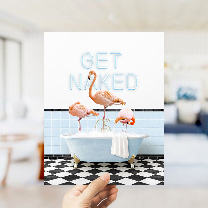 A hand holding a framed print with a vibrant image of three flamingos standing in a bathtub, with the words "GET NAKED" written on the wall behind them.