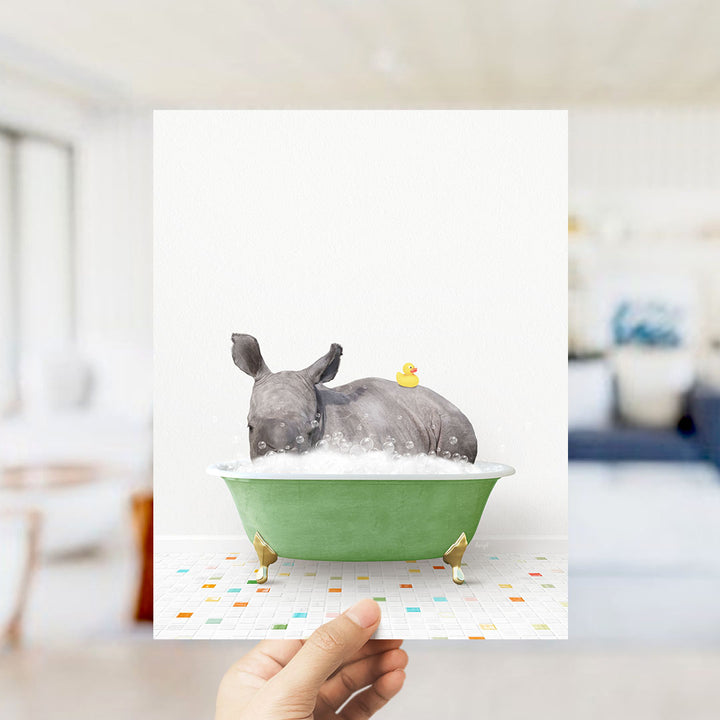 A hand holds up a framed image depicting a rhinoceros sitting in a green bathtub filled with bubbles, with a yellow rubber duck floating on the water.