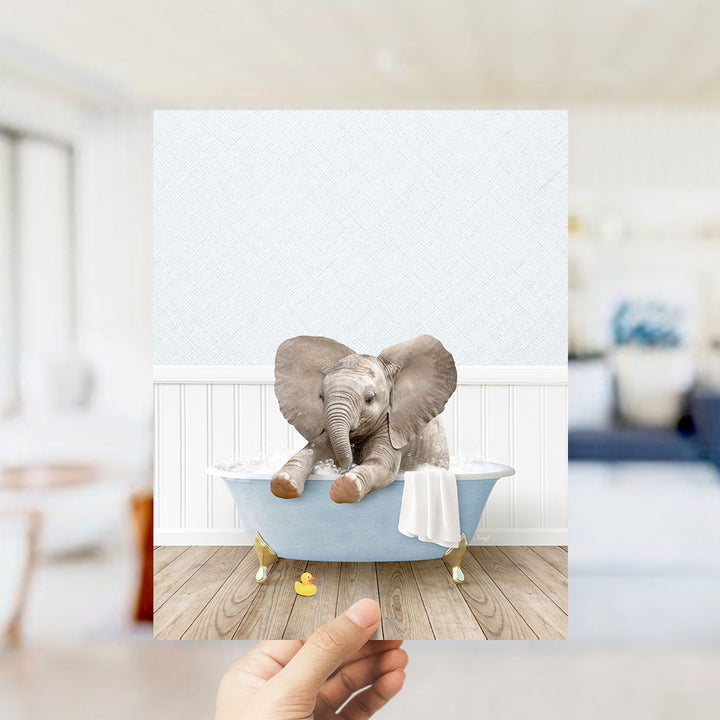 A hand holds up a framed image of a baby elephant sitting in a bathtub, surrounded by a white bathtub, a towel, and a rubber duck.