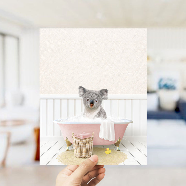 A hand holds up a framed image of a koala sitting in a pink bathtub, surrounded by a yellow rubber duck and a white towel.