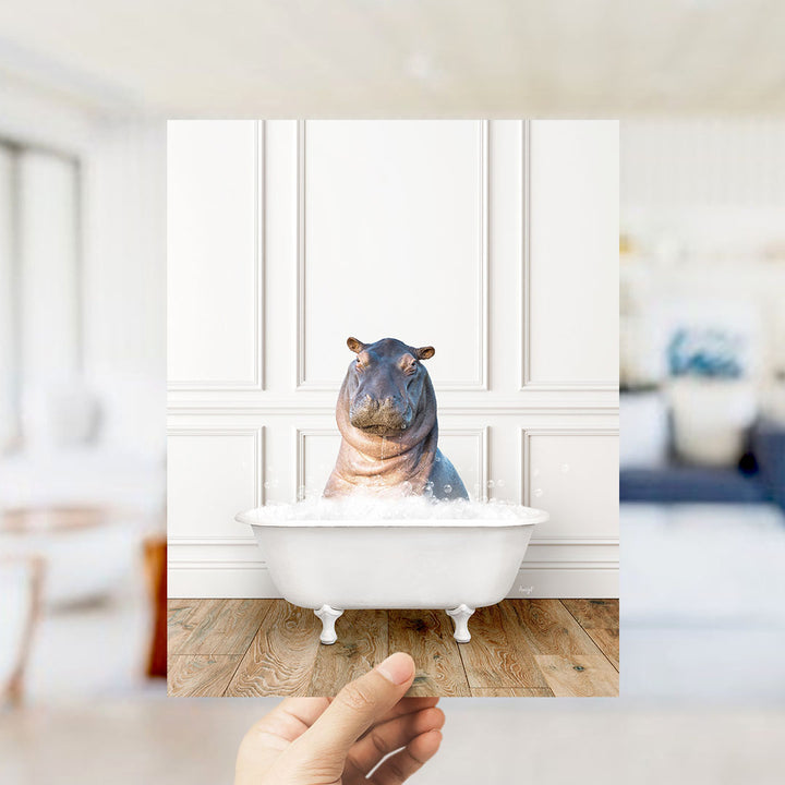 A hand holds up a framed image of a hippopotamus sitting in a bathtub filled with water, creating a whimsical and playful scene.