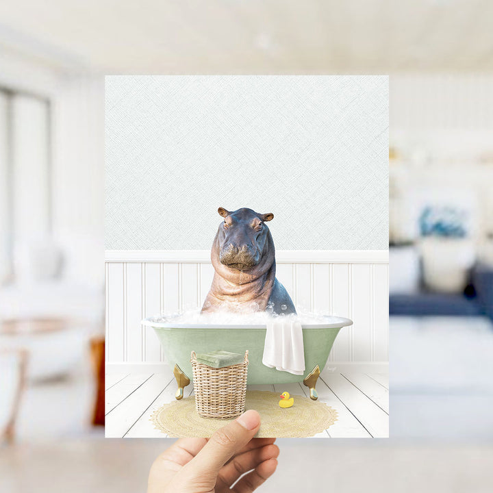 A hand holds up a framed image depicting a hippopotamus sitting in a bathtub filled with bubbles, surrounded by a yellow rubber duck and a white towel.