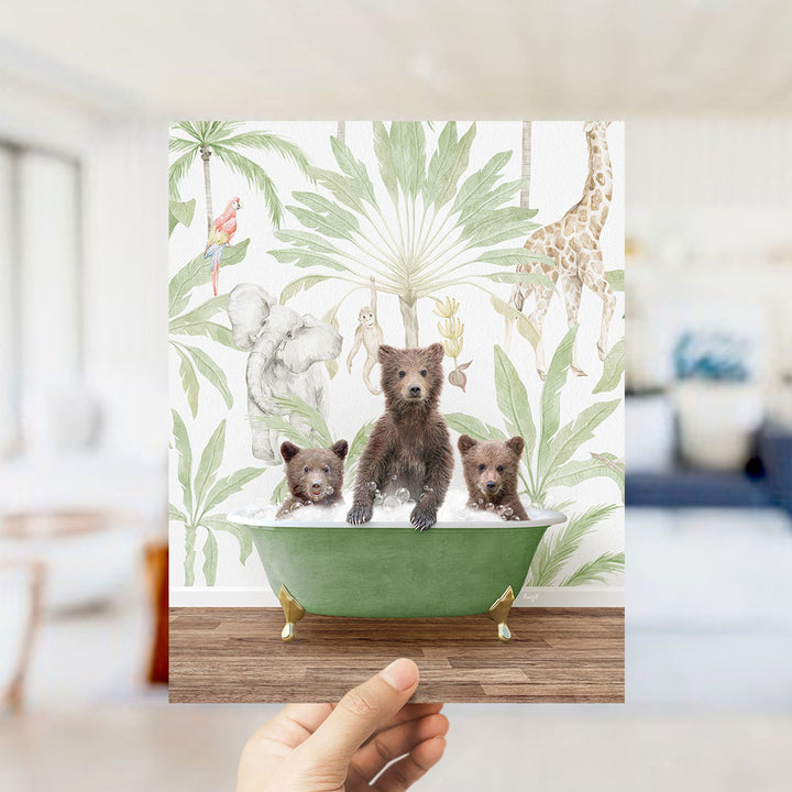 A hand holds up a framed print featuring a whimsical scene of three bears sitting in a green bathtub, surrounded by tropical foliage and animals.