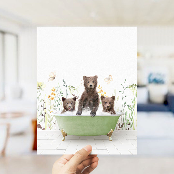 A hand is holding up a framed image of three bears sitting in a green bathtub, surrounded by flowers and butterflies.