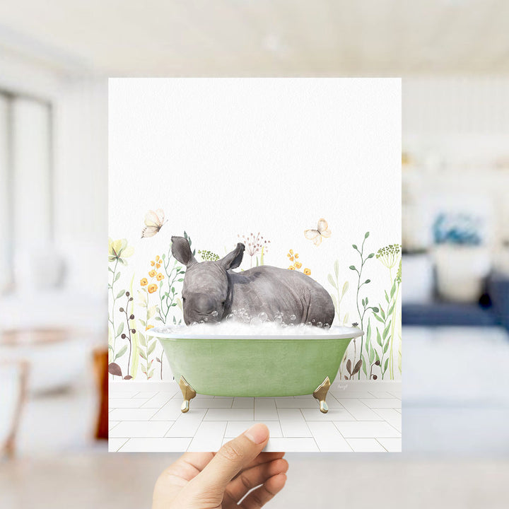 A hand holds up a framed image depicting a rhinoceros sitting in a green bathtub filled with bubbles, surrounded by a floral and butterfly-themed background.