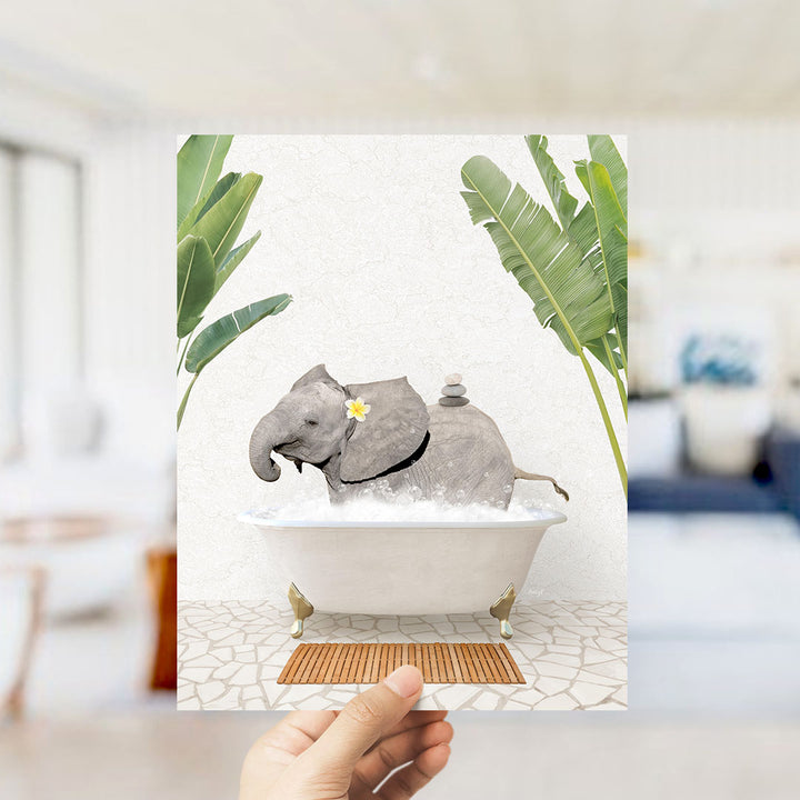 A hand is holding up a framed image depicting a gray elephant sitting in a white bathtub, surrounded by green leaves.