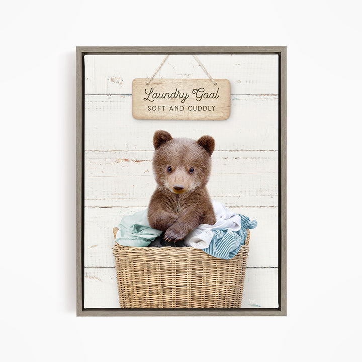 A cute brown bear sitting in a wicker laundry basket, with a wooden sign above that says "Laundry Goal Soft and Cuddly".