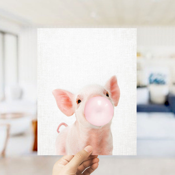 A hand is holding up a white square with a pink pig blowing a bubble with a pink bubble gum bubble.