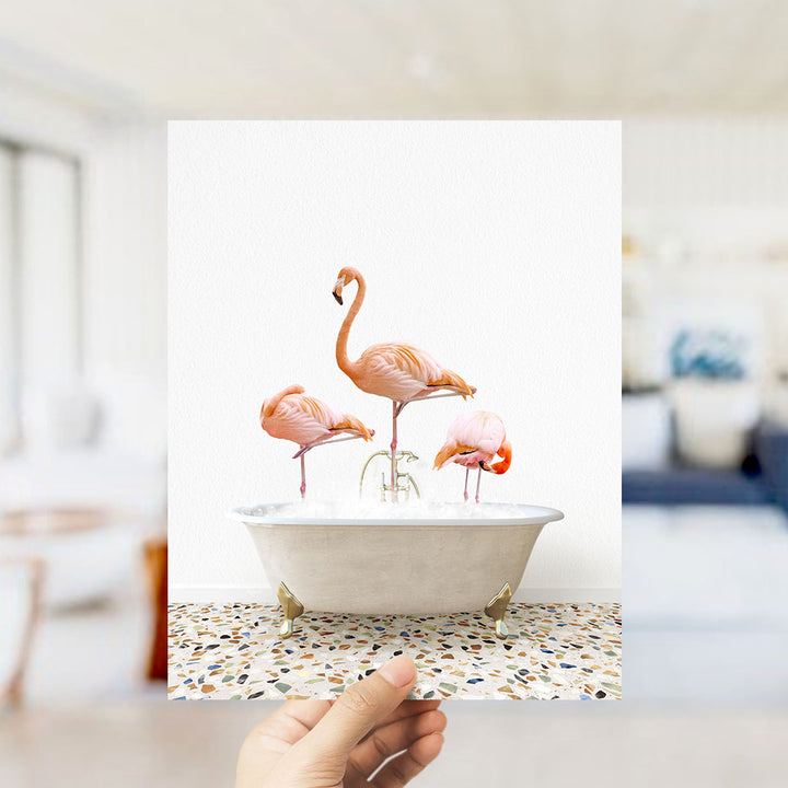 A hand holds up a framed photograph of three flamingos standing in a white bathtub filled with water.