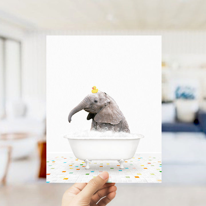 A hand holds up a framed image depicting a baby elephant sitting in a bathtub filled with bubbles, with a yellow rubber duck floating on its head.