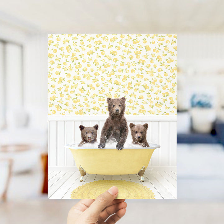 A hand holds up a framed image depicting three adorable brown bears sitting in a yellow bathtub, surrounded by a yellow and white floral pattern.