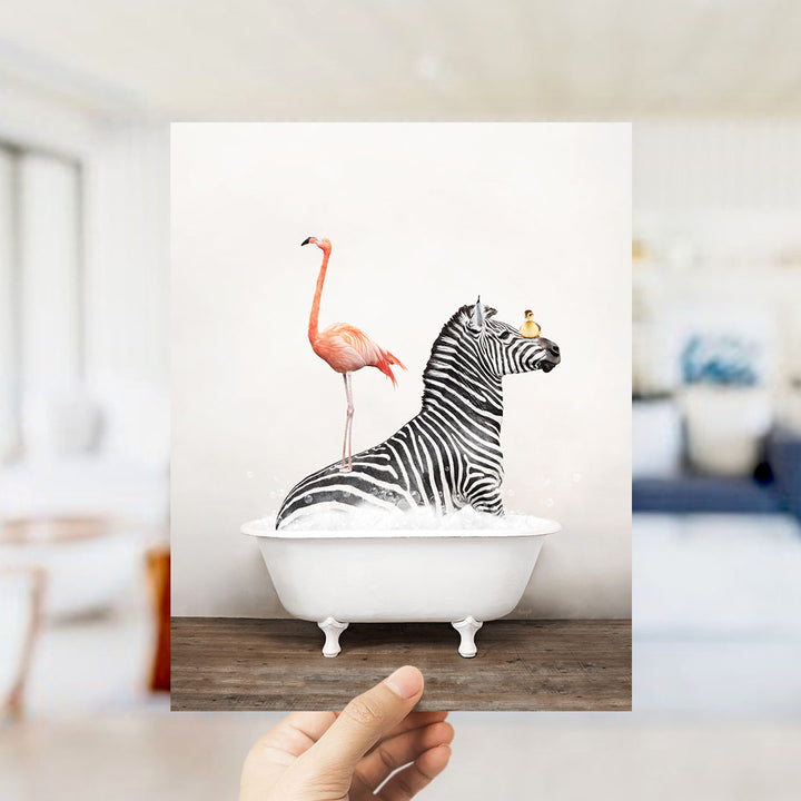 A hand holds up a framed image depicting a zebra standing in a bathtub with a pink flamingo.