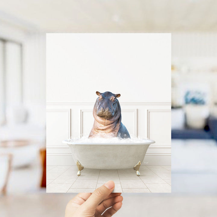 A hand holds up a framed image of a hippopotamus sitting in a bathtub filled with water, creating a whimsical and playful scene.