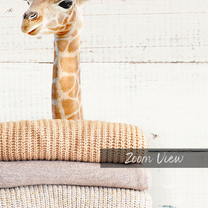 A giraffe peeking out from behind a stack of beige and brown knitted blankets, with the text "Zoom View" overlaid on the image.