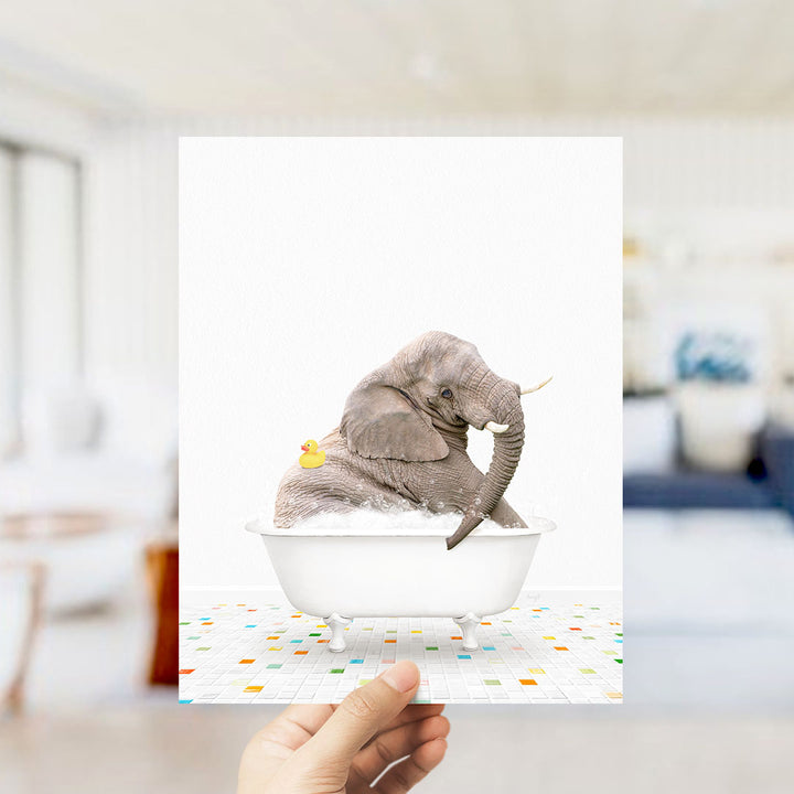 A hand holds up a framed image depicting a gray elephant sitting in a white bathtub, with a yellow rubber duck floating on its back.