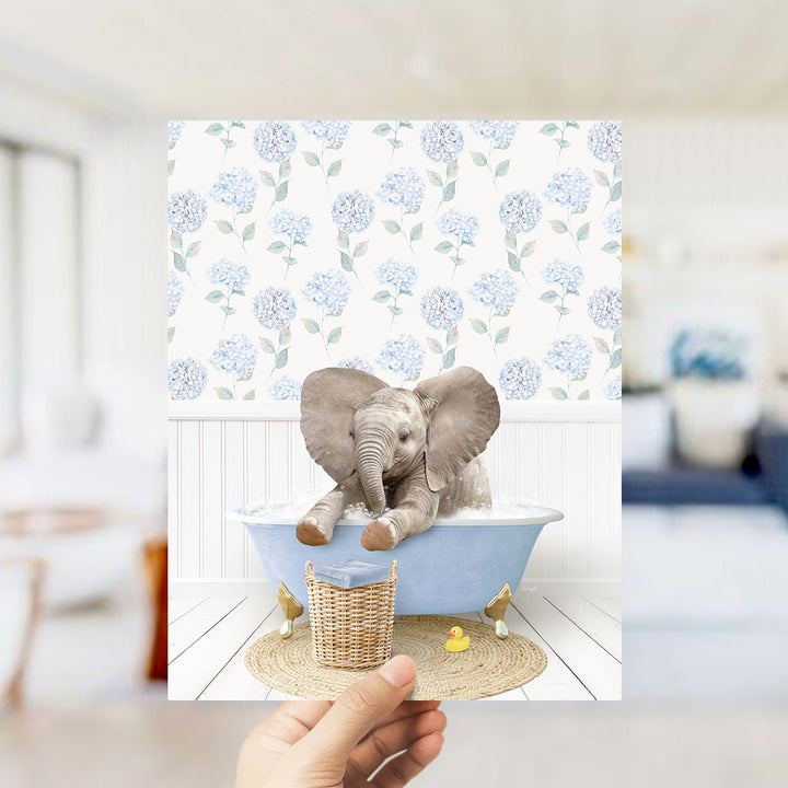 A hand holds up a framed image depicting a baby elephant sitting in a blue bathtub, surrounded by a floral patterned wall and a yellow rubber duck.