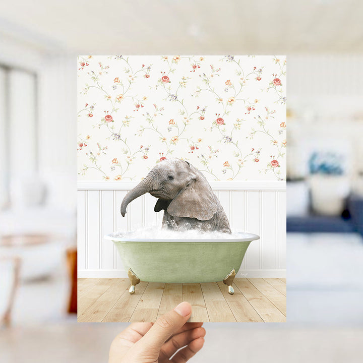 A hand holds up a framed image of a baby elephant sitting in a green bathtub filled with water, surrounded by a floral wallpaper.