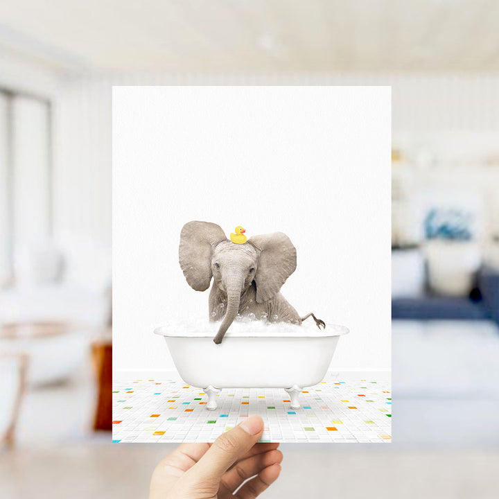 A hand holds up a framed image of a baby elephant sitting in a bathtub, with a yellow rubber duck on its head.