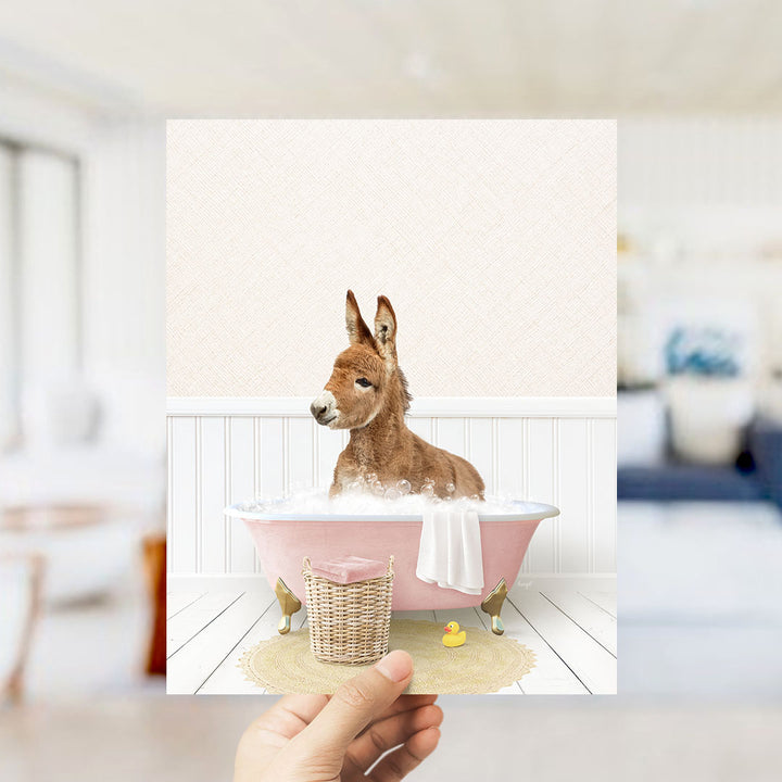 A donkey is sitting in a pink bathtub filled with bubbles, surrounded by a yellow rubber duck and a white towel.
