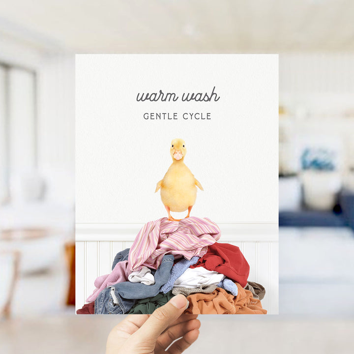 A hand holding a card with a yellow duck standing on a pile of laundry.