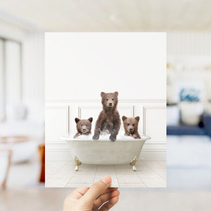 A hand holds up a framed image of three adorable brown bears sitting in a bathtub filled with water, creating a playful and whimsical scene.