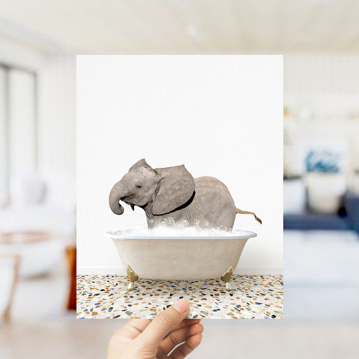 A hand holds up a framed image of a baby elephant sitting in a bathtub filled with water, creating a whimsical and playful scene.