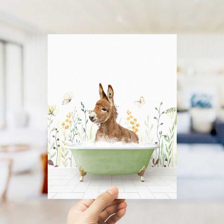 A hand holds up a framed image depicting a donkey sitting in a green bathtub filled with bubbles, surrounded by a floral background.