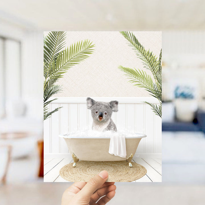 A hand holds up a framed image depicting a koala sitting in a bathtub filled with bubbles, surrounded by palm leaves.