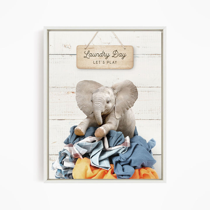 A framed image of a baby elephant sitting atop a pile of laundry, with a wooden sign above that reads "Laundry Day Let's Play".