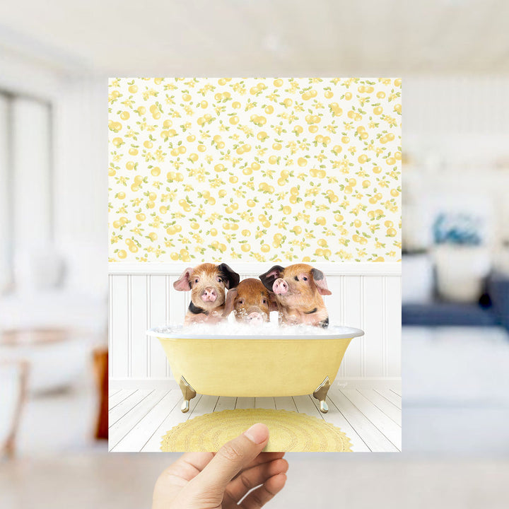 A hand holds up a framed image of three adorable pigs sitting in a yellow bathtub, surrounded by a yellow and white floral pattern.