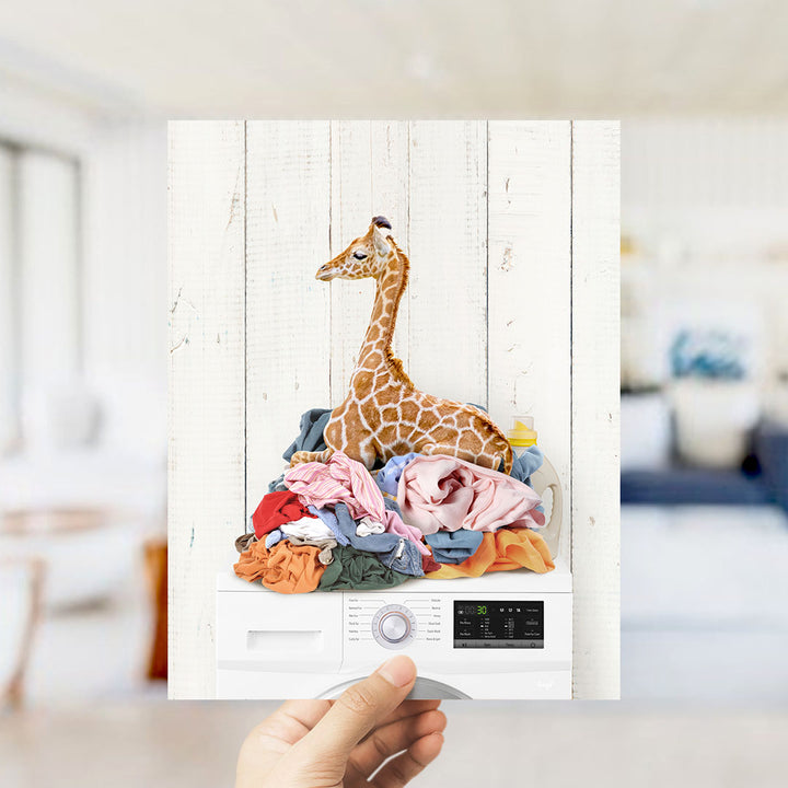 A hand holds up a photograph of a giraffe sitting atop a pile of laundry, with various clothing items surrounding it.