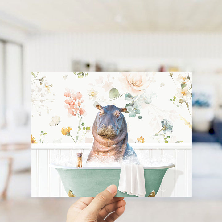 A hand holding a framed print featuring a hippopotamus sitting in a bathtub filled with water and flowers.