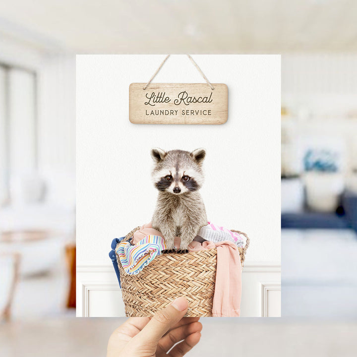 A raccoon sitting in a woven laundry basket, with a wooden sign above it that reads "Little Racal Laundry Service".