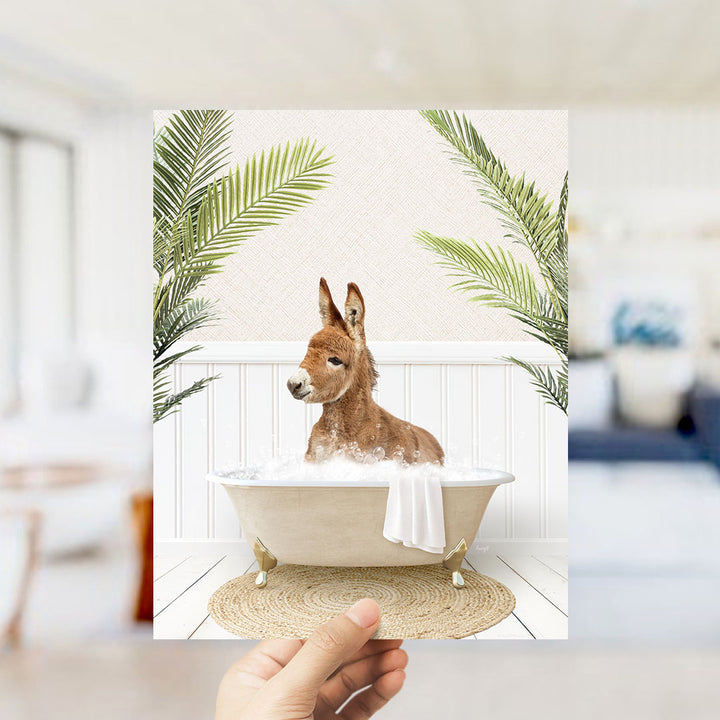 A hand holds up a framed image depicting a donkey sitting in a bathtub filled with bubbles, surrounded by palm leaves.