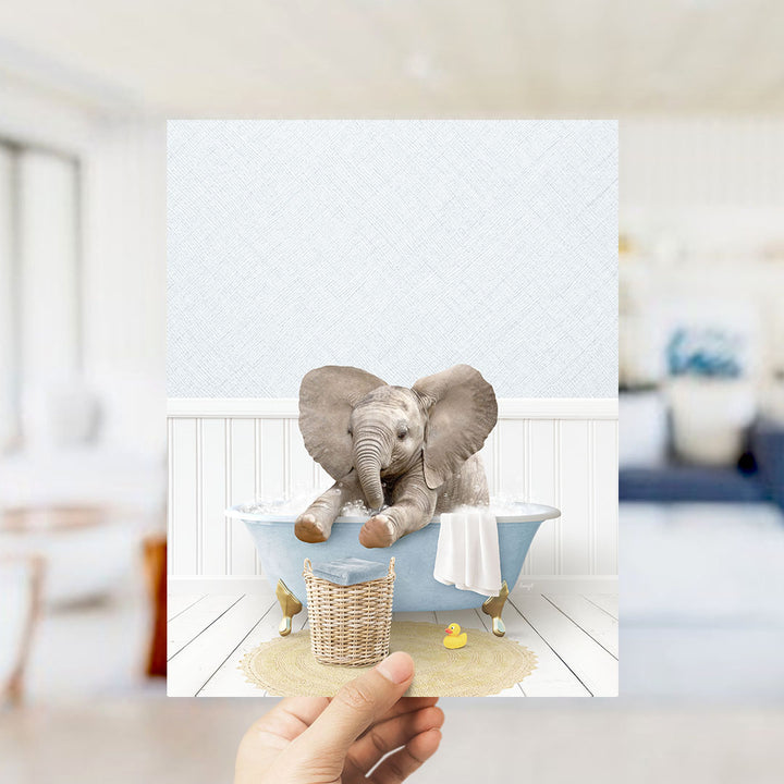 A hand holds up a framed image depicting a baby elephant sitting in a bathtub, surrounded by a basket of toys and a yellow rubber duck.