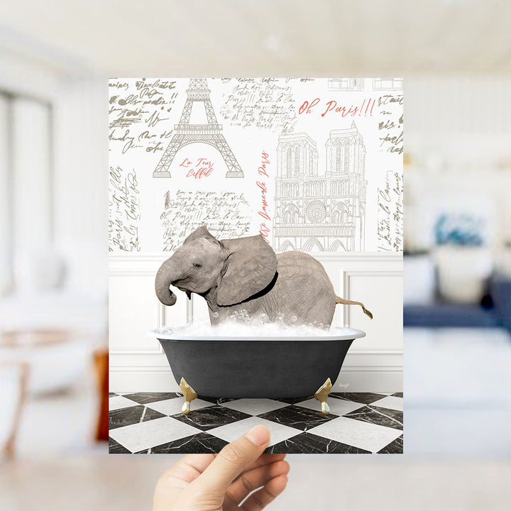 A hand holds up a printed image featuring a gray elephant sitting in a bathtub, surrounded by various Paris-themed elements such as the Eiffel Tower and other landmarks.