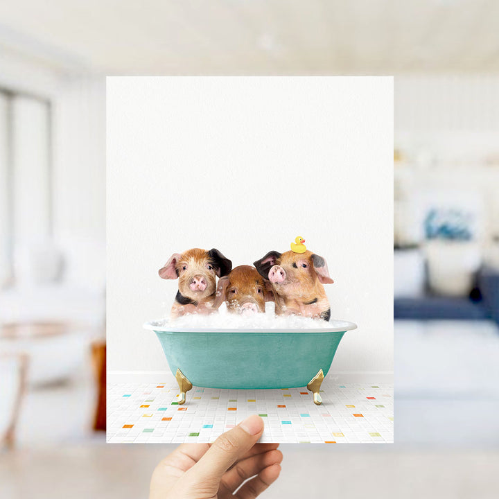 A hand holds up a framed image of three adorable pigs sitting in a green bathtub filled with bubbles, with a yellow rubber duck perched on the side.