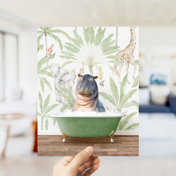 A hand holds up a framed image depicting a hippopotamus sitting in a green bathtub surrounded by tropical foliage, including a giraffe and palm trees.