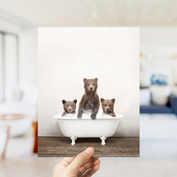 A hand holds up a framed image showing three baby bears sitting in a bathtub.