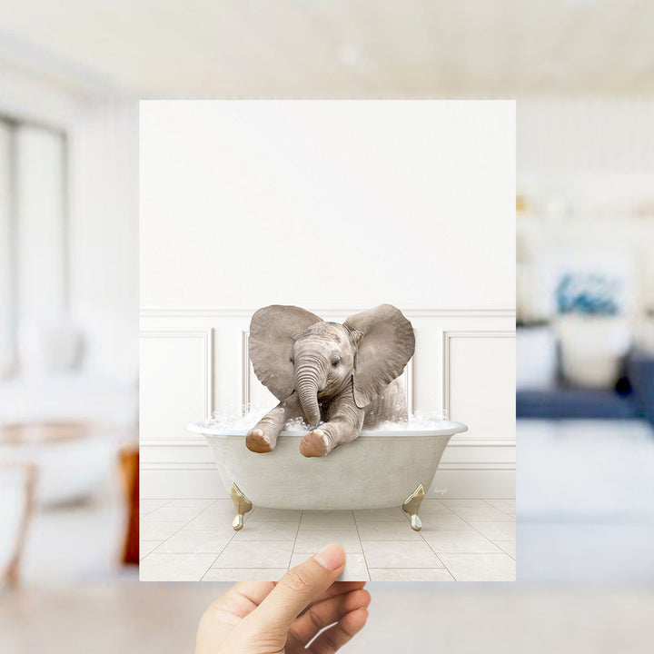 A hand holds up a framed image of a baby elephant sitting in a bathtub filled with water, creating a whimsical and playful scene.