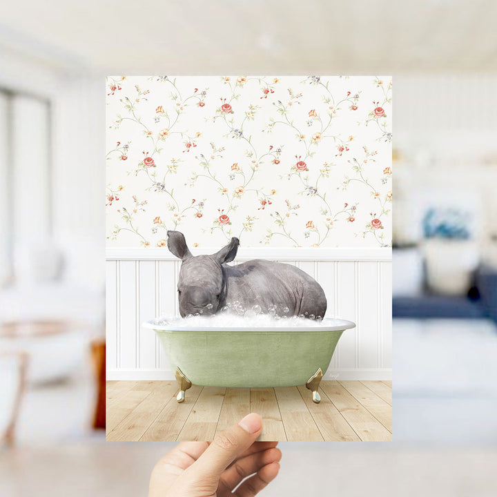 A hand holds up a framed image depicting a rhinoceros sitting in a green bathtub filled with bubbles, set against a floral wallpaper backdrop.