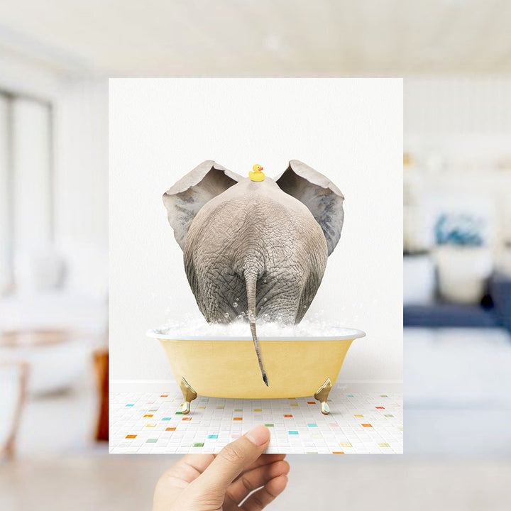 A hand holds up a framed image depicting an elephant sitting in a yellow bathtub, with a rubber duck perched on its head.