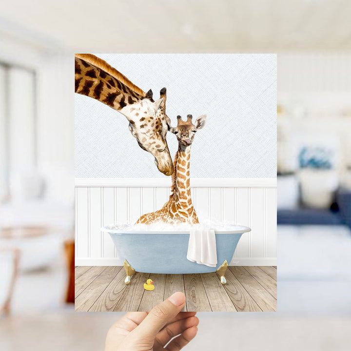 A hand holds up a framed image depicting two giraffes in a bathtub, with one giraffe sitting in the tub and the other standing nearby.