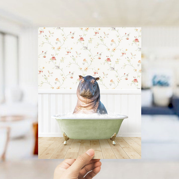 A hand holds up a framed image of a hippopotamus sitting in a bathtub filled with bubbles, against a floral wallpaper backdrop.