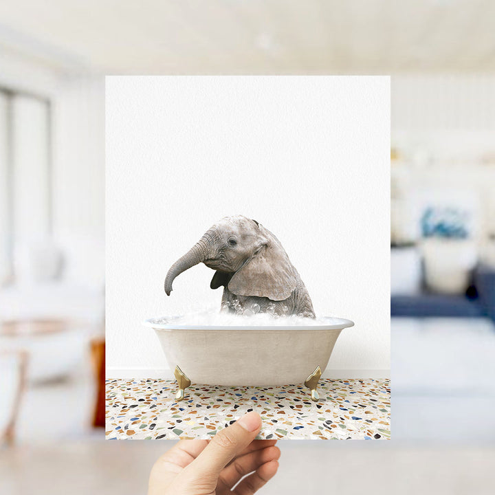 A hand holds up a framed image of a baby elephant sitting in a bathtub filled with water, creating a whimsical and playful scene.