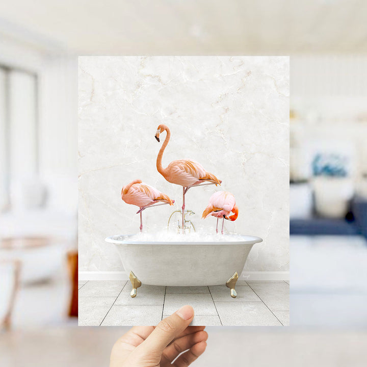 A hand holds up a framed image of three flamingos standing in a white bathtub filled with water.