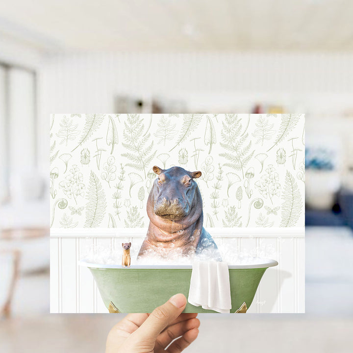A hand holds up a framed image depicting a hippopotamus sitting in a bathtub, surrounded by a green tub and a small dog.